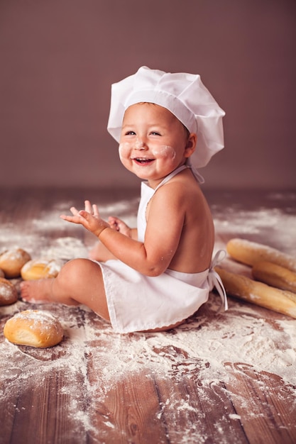 Charming little girl in hat of cook and apron sitting in flour with bread loaves laughing happily