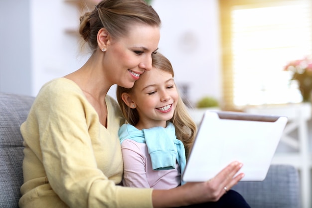 Charming little girl and her beautiful young mom using digital tablet and smiling while sitting on sofa at home