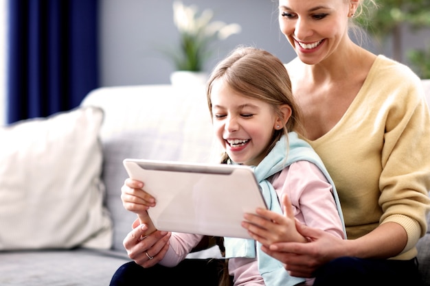 Charming little girl and her beautiful young mom using digital tablet and smiling while sitting on sofa at home