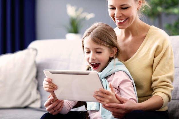 Charming little girl and her beautiful young mom using digital tablet and smiling while sitting on sofa at home