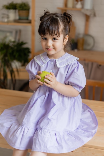 charming little girl in a lilac dress eats a green apple in the kitchen. space for text, banner