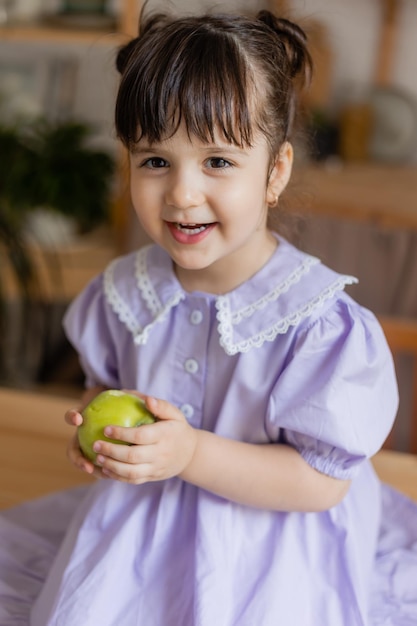 charming little girl in a lilac dress eats a green apple in the kitchen. space for text, banner