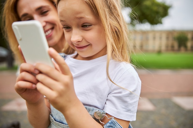 charming little girl looking on the screen cell phone while standing in front of adult woman