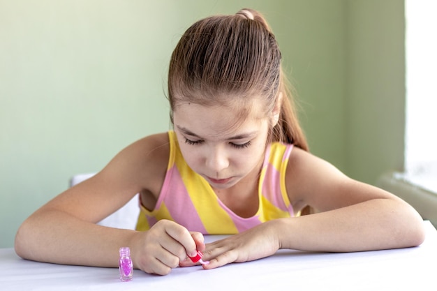 A charming little girl sits at the table and paints her fingers with nail polish