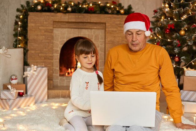 Charming little girl with grandfather sitting and using digital laptop during Christmas Eve, sitting on floor on soft carper near fir tree and fireplace, family looking concentrated at screen.