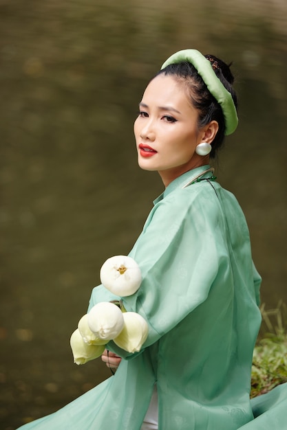 Photo charming lovely young woman in traditional vietnamese dress and headwear resting by water on summer day