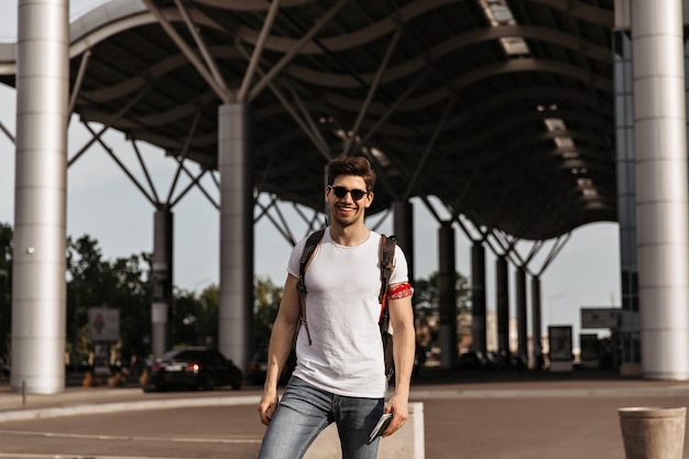 Charming man in white tee and jeans smiling Brunette guy in sunglasses holds passport and backpack and poses near airport