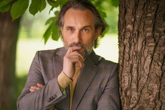 Charming mature grey birded businessman in casual standing under tree on the grass looking at camera with arms folded touching his face Freelancer spend time working outdoors