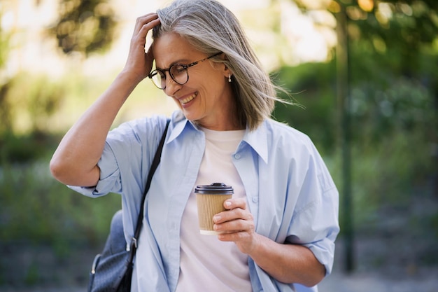 Charming mature woman drinks coffee on the go fixing her hair Mature woman spend free time traveling in european old town enjoying cup of tea coffee Grey haired woman in casual with backpack