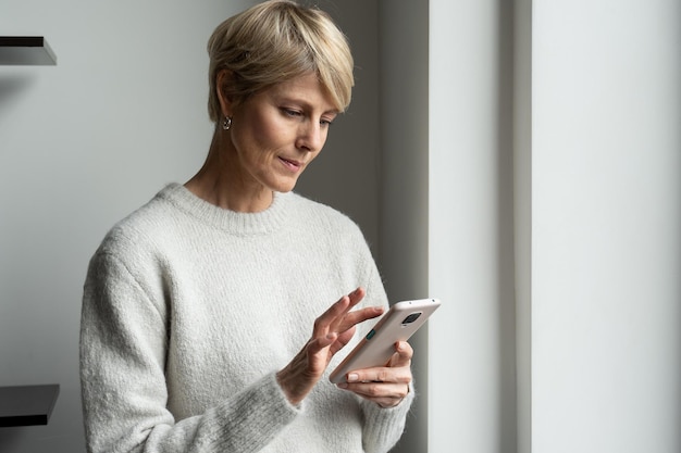 Charming middleaged woman standing at the window and typing text messages on her phone using a