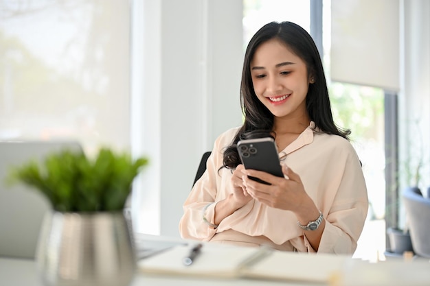 Charming millennial Asian businesswoman using her smartphone at her desk chat text