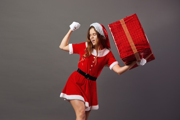 Charming mrs.Claus dressed in the red robe, Santa's hat and white gloves holds in her hand the huge Christmas present on the gray background .