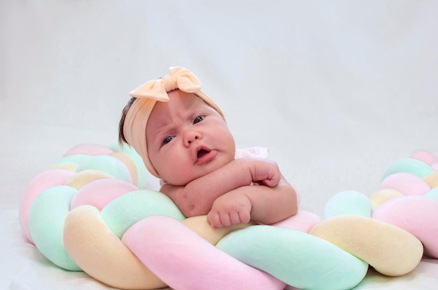 Charming newborn baby with bow on her head is lying on her stomach on a white bed at home looking into the camera morning of the child Concept of children's goods Happy childhood of healthy child