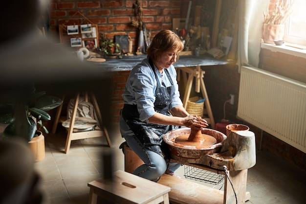 Charming old woman in apron sitting on bench and working on pottery wheel