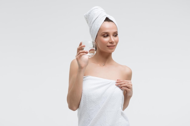 Charming pleasant woman in fluffy white towel on head with glass of clean water looking away