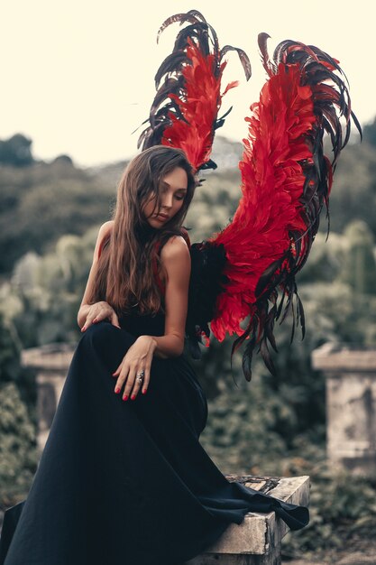 Charming portrait of dark angel  with red strong powerful wings in black lace dress  and with cold eyes, art photo. Outdoor photo