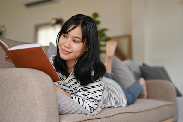 Charming and relaxed Asian female enjoys reading a book while lying on her comfortable sofa