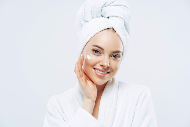 Charming relaxed beauty woman applies face cream, cares about complexion, touches cheek with hand, smiles gently dressed in bath robe, wrapped towel on washed hair, isolated on white