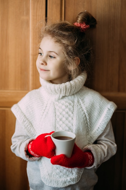 Charming seven-year-old girl drinks warm tea, standing next to the window. She is wearing a warm knitted sweater, her hands are in red knitted gloves. Christmas time.