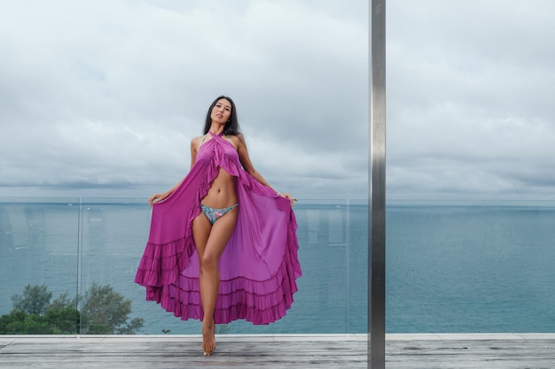 Charming slender brunette in a purple cape posing while standing on the terrace against the backdrop of the seascape