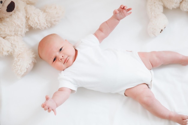 A charming smiling blueeyed 2monthold baby in a white bodysuit lies in a crib next to a teddy bear View from above a newborn