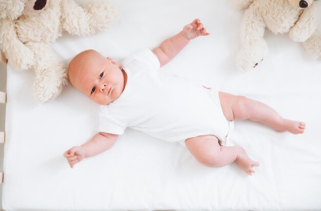 Photo a charming smiling blueeyed 2monthold baby in a white bodysuit lies in a crib next to a teddy bear view from above a newborn