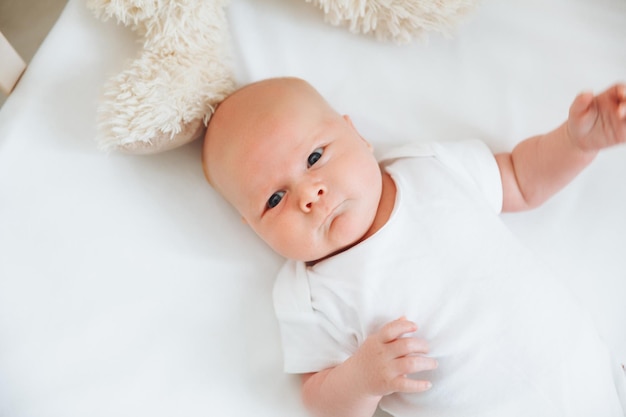 A charming smiling blueeyed 2monthold baby in a white bodysuit lies in a crib next to a teddy bear View from above a newborn