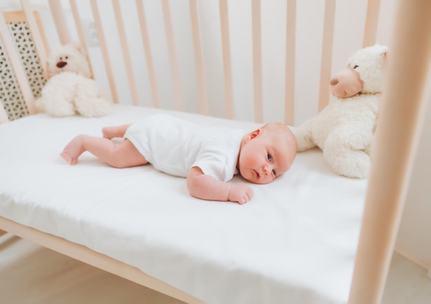 A charming smiling blueeyed 2monthold baby in a white bodysuit lies in a crib next to a teddy bear View from above a newborn