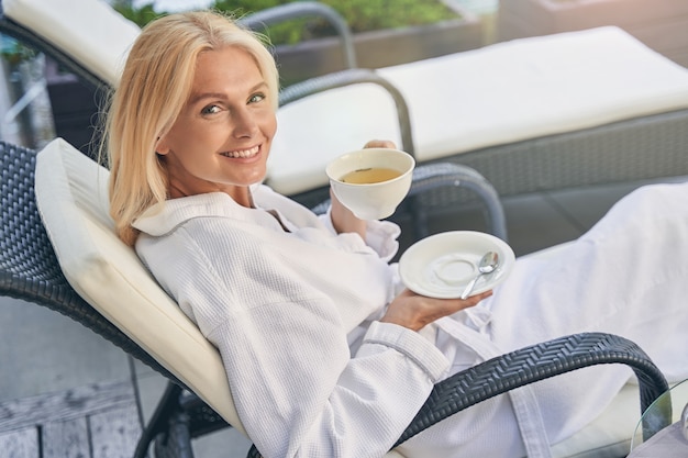 charming smiling female lying in the deckchair holding white cup in hand