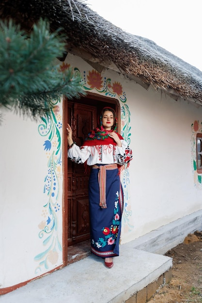 Charming smiling woman in traditional ukrainian handkerchief necklace and embroidered dress standing at background of decorated hut Ukraine style folk ethnic culture