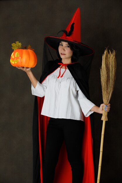 Charming smiling young Asian woman in witch costume with Halloween pumpkin and broom looking at camera against dark gray background