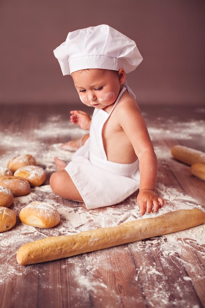 Charming toddler baby in hat of cook and apron sitting in flour with bread loaves playing with baguette