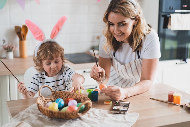 Charming woman and her twoyearold son in bunny ears paint Easter eggs with paints sitting at the table on the kitchen