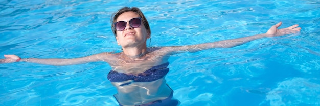 Charming woman in sunglasses enjoys swimming in turquoise water of pool