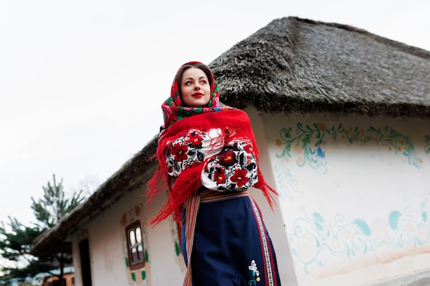 Charming woman in traditional ukrainian handkerchief necklace and embroidered dress standing at background of decorated hut Ukraine style folk ethnic culture