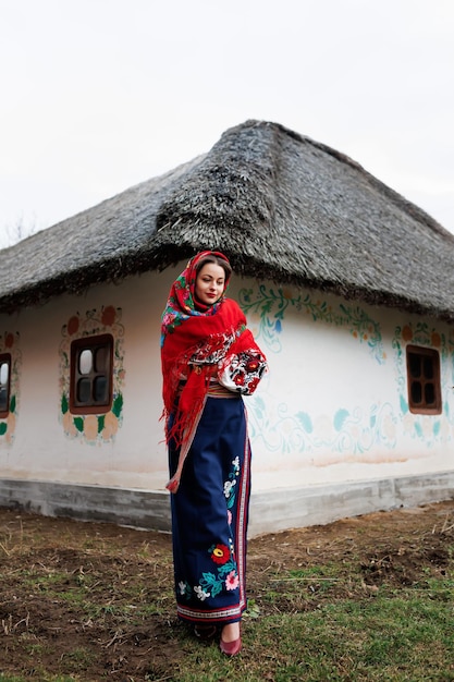 Charming woman in traditional ukrainian handkerchief necklace and embroidered dress standing at background of decorated hut Ukraine style folk ethnic culture