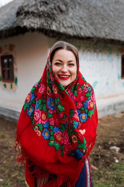 Charming woman in traditional ukrainian handkerchief necklace and embroidered dress standing at background of decorated hut with thatched roof Ukraine style folk ethnic culture