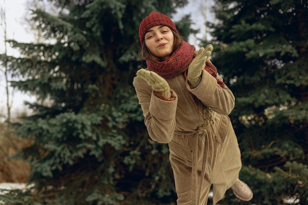 Charming woman in warm winter wear sending air kiss and looking at camera in park