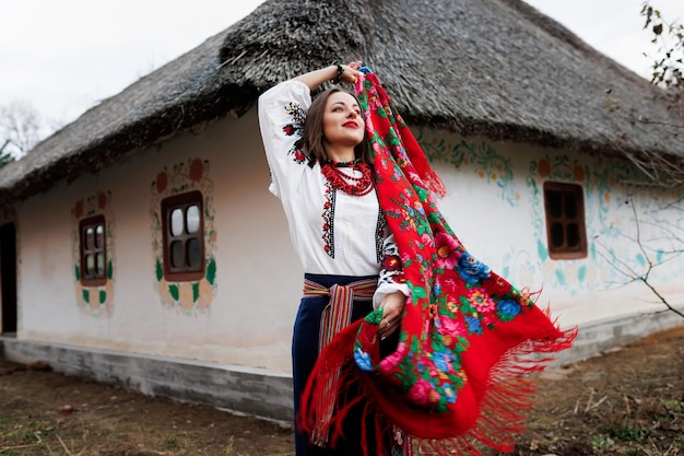 Charming woman with traditional ukrainian handkerchief necklace and embroidered dress standing at background of decorated hut with thatched roof Ukraine style folk ethnic culture