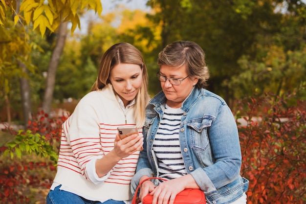 Charming women in casual style mom and daughter are sitting on bench in beautiful autumn park and looking at photos on phone