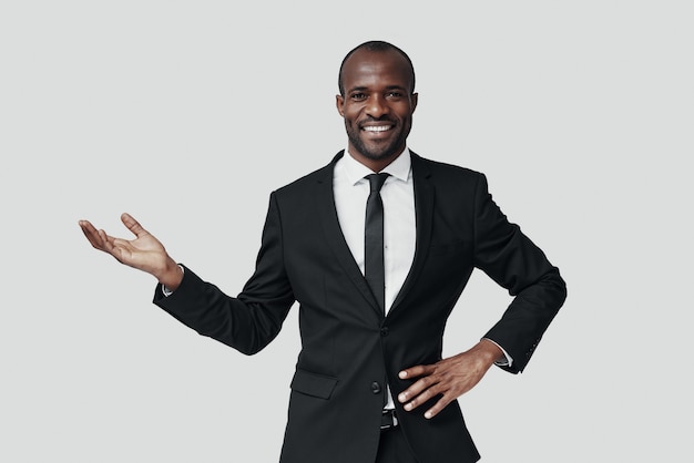 Charming young African man in formalwear pointing copy space and smiling while standing against grey wall