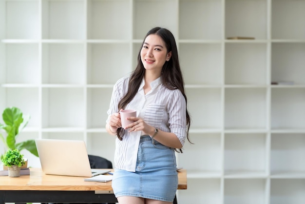 Charming young Asian businesswoman standing holding coffee mug at the office Looking at camera