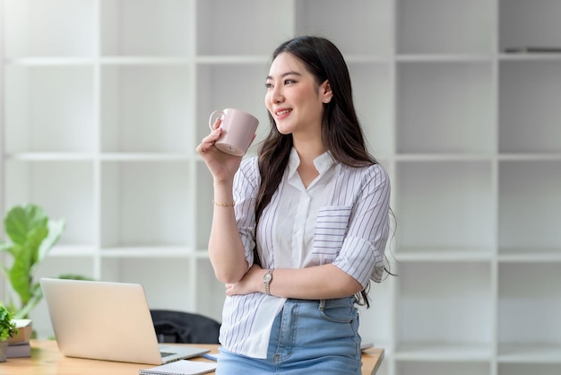 Charming young Asian businesswoman standing holding coffee mug at the office.