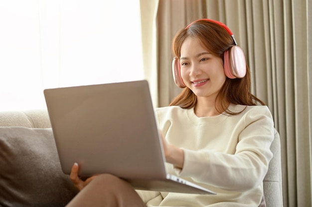 Charming young Asian female in living room wearing headphones and using laptop computer