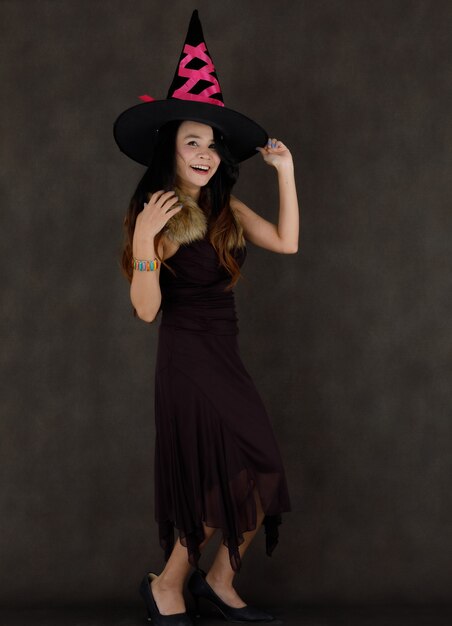 Photo charming young asian female in sorceress costume and hat smiling and looking at camera against dark gray background