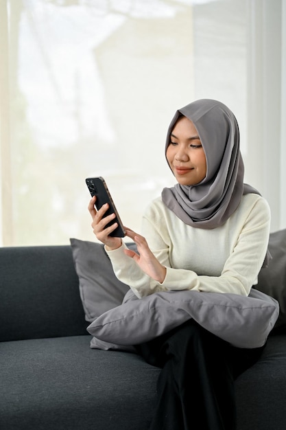 Charming young Asian Muslim woman wearing hijab using her smartphone while relaxing on sofa