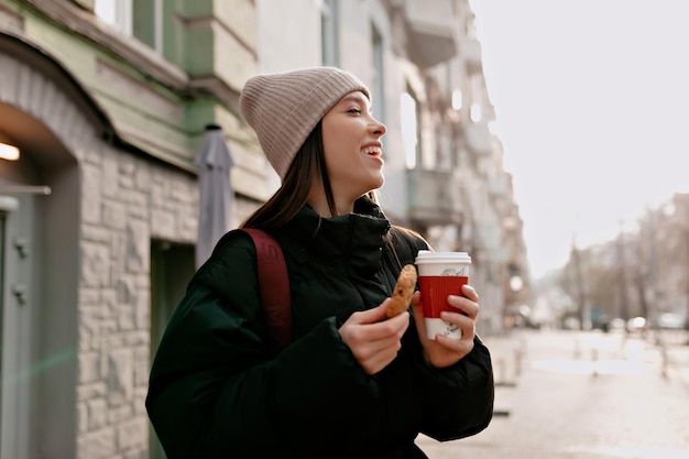 Charming young brunette woman in beige hat dark jacket is smiling and looking at tasty cookies while walking on sunny city street