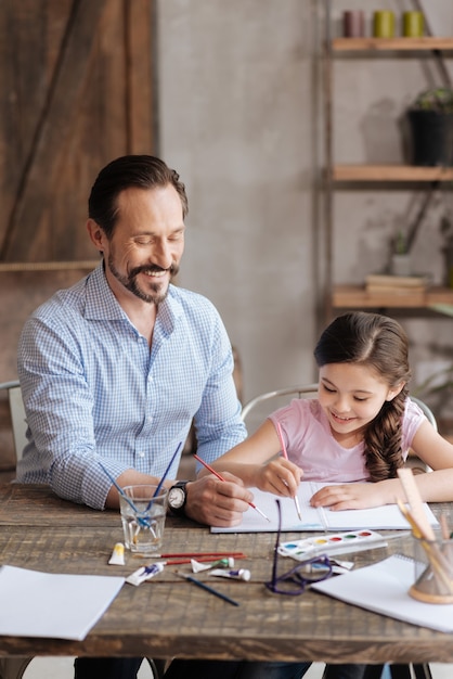 Charming young father and his petite pleasant daughter sitting at the table and painting a picture with watercolors, being happy to do it together