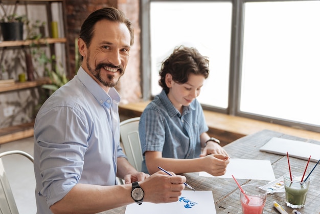 Charming young father painting a watercolor picture together with his son while looking pleased to spend some time with him