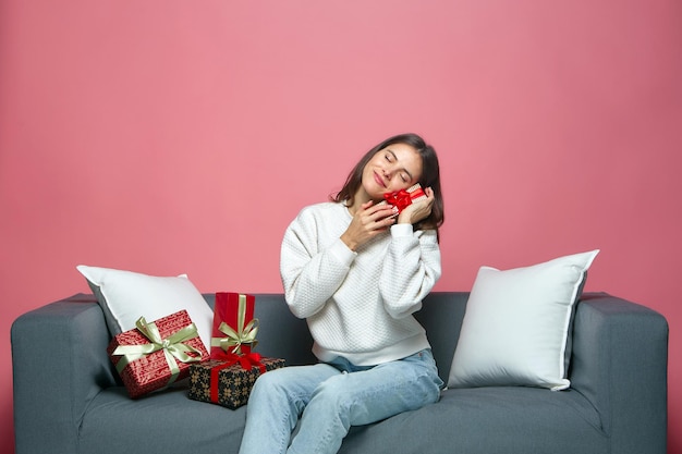 Charming young girl holding gift in beautiful package sitting on sofa celebrating christmas or